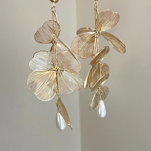 以下が含まれることがあります： 金色の花のドロップイヤリング。各イヤリングは、複数の繊細な、重ねられた花の形と、真珠色の涙型のビーズが特徴です。イヤリングはポストバッククロージャーと上部に小さな金のボールが付いています。