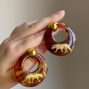 Peut inclure: Une paire de grandes boucles d'oreilles créoles avec un motif écaille de tortue. Chaque boucle d'oreille présente un charm de tigre doré et un clou doré en haut. Les boucles d'oreilles sont tenues par une main.
