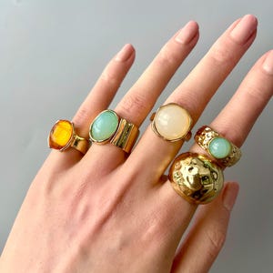 May include: A hand displays six gold-toned rings with various gemstone settings. The rings feature amber, turquoise, and cream-colored stones, as well as textured and smooth gold bands. The rings are in different shapes and sizes.