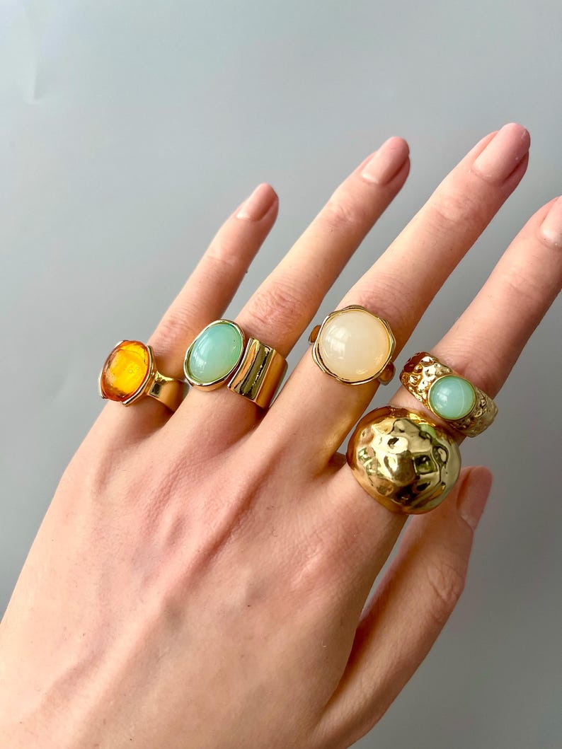 May include: A hand displays six gold-toned rings with various gemstone settings. The rings feature amber, turquoise, and cream-colored stones, as well as textured and smooth gold bands. The rings are in different shapes and sizes.