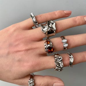 May include: A collection of silver rings with unique, textured designs. Some rings feature smooth, oval-shaped white stones, while others have an organic, molten metal appearance. One ring showcases a large, amber-colored stone.