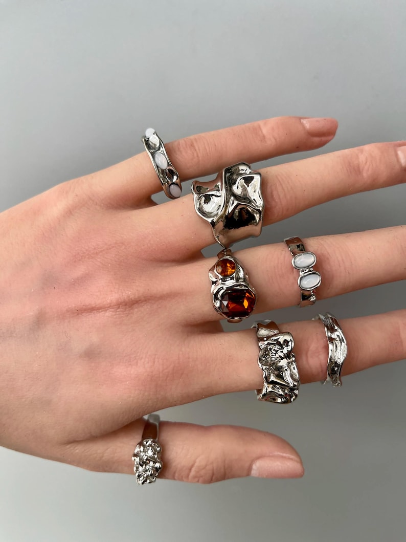 May include: A collection of silver rings with unique, textured designs. Some rings feature smooth, oval-shaped white stones, while others have an organic, molten metal appearance. One ring showcases a large, amber-colored stone.