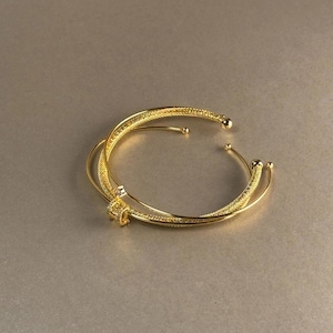 Può includere: Due braccialetti color oro sono intrecciati, creando un effetto a strati. I braccialetti hanno un design strutturato e una finitura liscia e lucida. I braccialetti sono aperti, con punte arrotondate. Un piccolo elemento decorativo collega i due braccialetti.