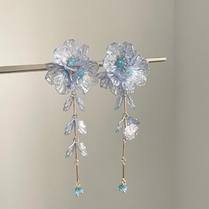 Puede incluir: Un par de pendientes florales azul claro con detalles dorados. Cada pendiente presenta una flor grande con detalles turquesa, elementos en forma de hojas en cascada y pequeñas cuentas turquesas en la parte inferior.