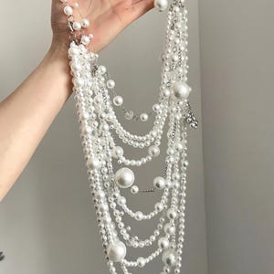 Può includere: Una collana di perle multifilo con perle di diverse dimensioni. La collana è realizzata con una catena d'argento e presenta perle grandi e rotonde.