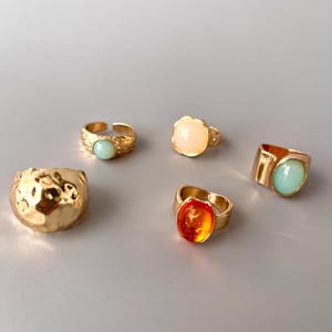 May include: A collection of five gold-toned rings. The rings feature various designs, including textured bands and settings with light blue, orange, and peach-colored stones. The rings are displayed on a light gray surface.