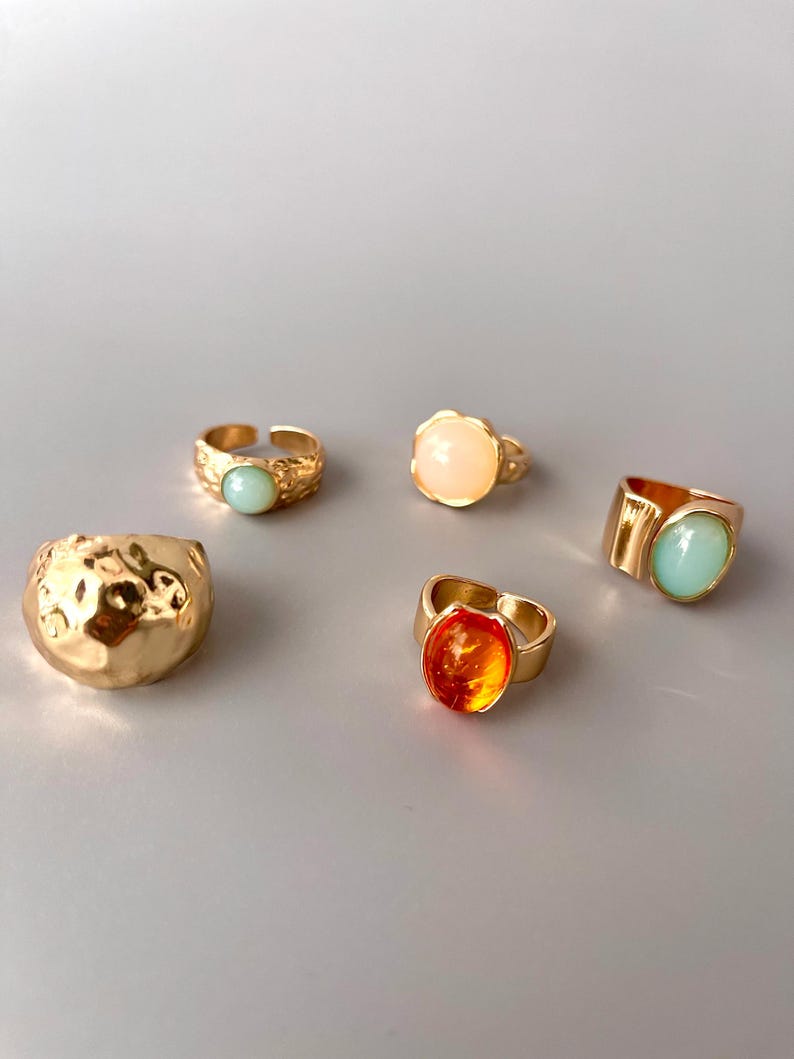 May include: A collection of five gold-toned rings. The rings feature various designs, including textured bands and settings with light blue, orange, and peach-colored stones. The rings are displayed on a light gray surface.