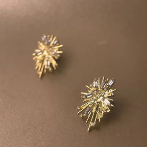 Może przedstawiać: Para złotych kolczyków sztyftów. Każdy kolczyk ma wzór rozbłysku gwiazdy z przezroczystymi, prostokątnymi i okrągłymi kryształami. Kolczyki są umieszczone na neutralnym, brązowym tle.