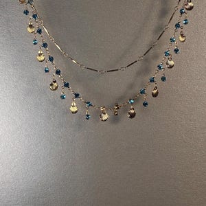 Puede incluir: Un collar de varias capas con cadenas doradas. El collar está adornado con pequeños colgantes de conchas doradas y cuentas de cristal azules. El diseño es delicado y adecuado para diversas ocasiones.