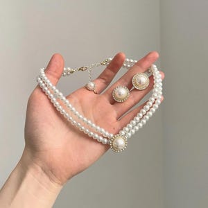 May include: A two-strand pearl necklace with a large pearl pendant and matching pearl earrings. The necklace and earrings feature a gold-colored setting with small, clear crystals around the pearls. The jewelry is displayed against a neutral background.