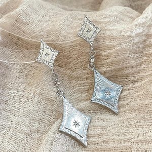 Puede incluir: Un par de pendientes plateados. Cada pendiente presenta un colgante en forma de diamante con un diseño de estrella en el centro. Los colgantes están conectados a una pieza superior cuadrada más pequeña con un diseño similar.