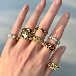 May include: A hand displaying a collection of gold-toned rings. The rings feature various designs, including textured, gemstone-set, and band styles. The rings are in different sizes and shapes, showcasing a variety of jewelry designs.