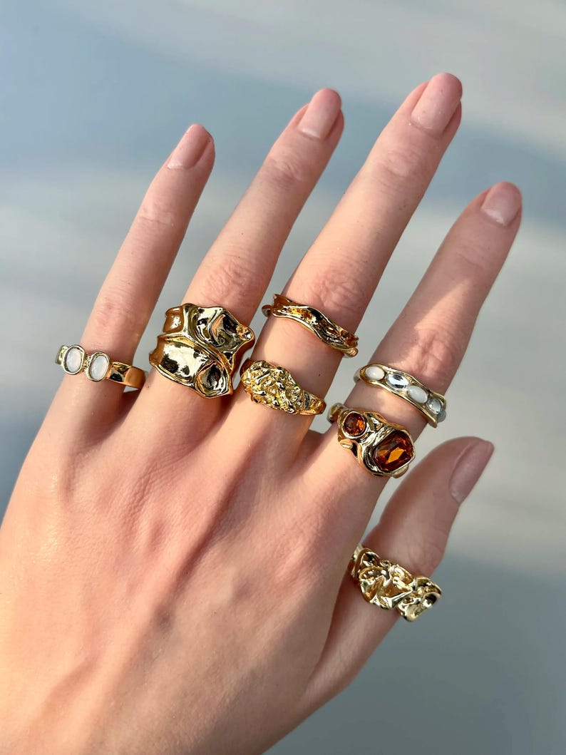 May include: A hand displaying a collection of gold-toned rings. The rings feature various designs, including textured, gemstone-set, and band styles. The rings are in different sizes and shapes, showcasing a variety of jewelry designs.