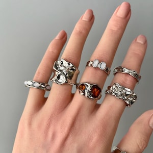May include: A hand displays a collection of silver rings with unique, textured designs. Some rings feature white or amber-colored stones, while others have abstract, molten metal-like shapes. The rings vary in size and style, showcasing a modern jewelry aesthetic.