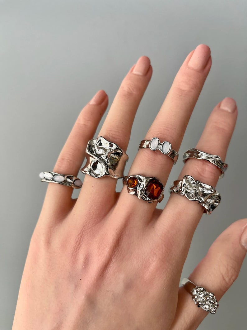 May include: A hand displays a collection of silver rings with unique, textured designs. Some rings feature white or amber-colored stones, while others have abstract, molten metal-like shapes. The rings vary in size and style, showcasing a modern jewelry aesthetic.