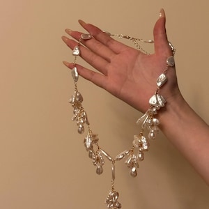 Op de afbeelding: Een goudkleurige ketting met onregelmatig gevormde witte parels. De ketting heeft een delicate ketting en een hanger met meerdere parelclusters, waardoor een watervaleffect ontstaat. De parels variëren in grootte en vorm.