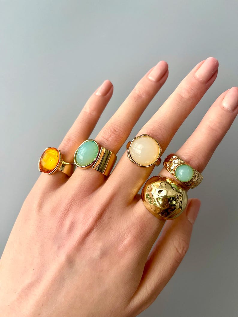 May include: A hand displays five gold-toned rings with various gemstone settings. The rings feature an amber-colored oval stone, a light green oval stone, a white oval stone, a textured gold band with a light green stone, and a large, textured gold dome ring.