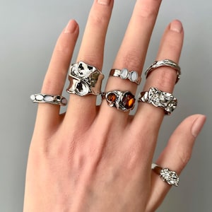 May include: A hand showing a selection of silver rings. The rings have diverse designs, including organic forms, gemstones, and textured finishes. The rings are presented against a neutral grey backdrop.