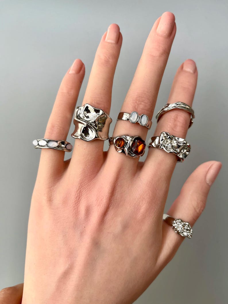 May include: A hand displaying a collection of silver rings. The rings feature various designs, including organic shapes, gemstones, and textured surfaces. The rings are displayed against a neutral gray background.