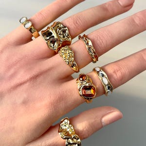 May include: A hand displays a collection of gold-toned rings. The rings feature various designs, including organic shapes, textured surfaces, and gemstones in shades of orange and white. The rings are displayed against a neutral background.