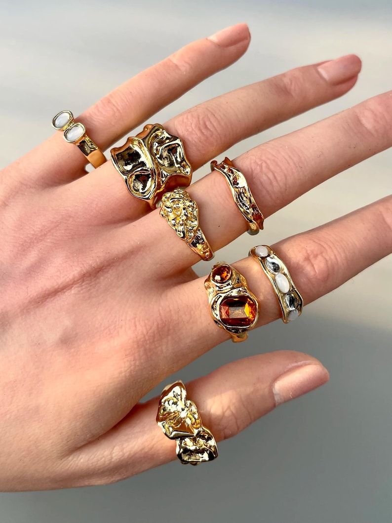 May include: A hand displays a collection of gold-toned rings. The rings feature various designs, including organic shapes, textured surfaces, and gemstones in shades of orange and white. The rings are displayed against a neutral background.