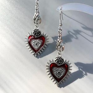 Puede incluir: Pendientes colgantes plateados. Cada pendiente presenta un colgante en forma de corazón rojo con un diseño de ojo, rodeado por un diseño de sol. También hay una rosa y elementos decorativos. Para orejas perforadas.