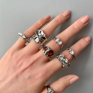 May include: A hand displaying a collection of silver rings. The rings feature various designs, including organic shapes, textured surfaces, and gemstone accents in shades of red and white. The rings are displayed against a neutral gray background.