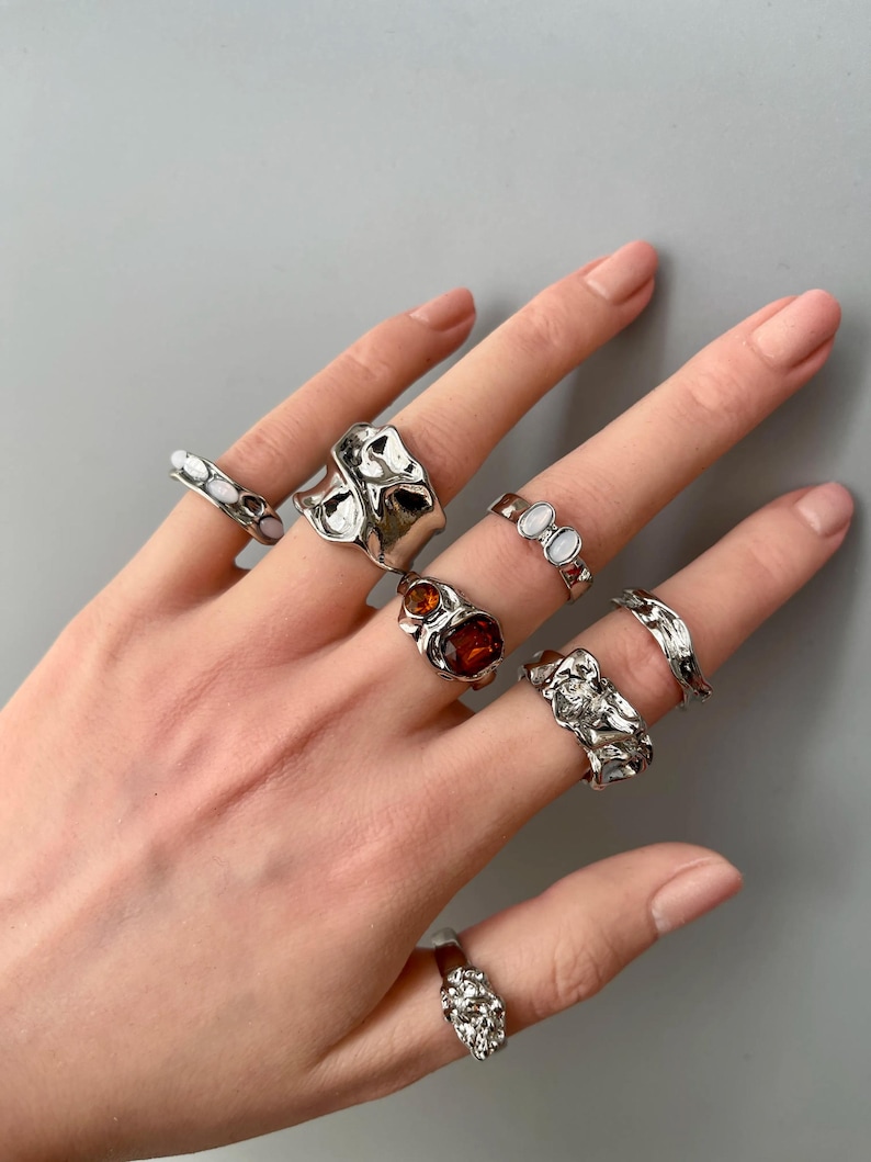 May include: A hand displaying a collection of silver rings. The rings feature various designs, including organic shapes, textured surfaces, and gemstone accents in shades of red and white. The rings are displayed against a neutral gray background.