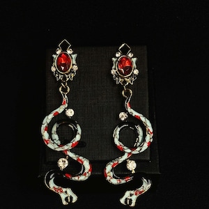 May include: A pair of gold-toned snake earrings with red and white enamel detailing. Each earring features a large, oval, red gemstone at the top, with smaller clear gemstones and a snake design that coils downward.