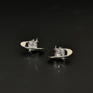 Puede incluir: Un par de pendientes de botón plateados. Cada pendiente presenta una gema cuadrada y transparente engastada en un diseño en forma de media luna con detalles de estrellas. Los pendientes se muestran sobre un fondo oscuro.