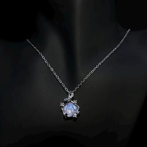 May include: A silver necklace featuring a seashell pendant with a shimmering blue center. The pendant is accented with small pink gemstones and a delicate chain. The necklace is displayed against a black background.