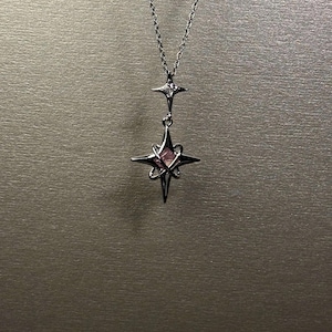 Pode incluir: Um colar prateado com um pendente em forma de estrela com uma pedra rosa no centro. O pendente é suspenso por uma corrente delicada, com uma estrela menor acima. O colar tem um design minimalista e elegante, adequado para qualquer ocasião.