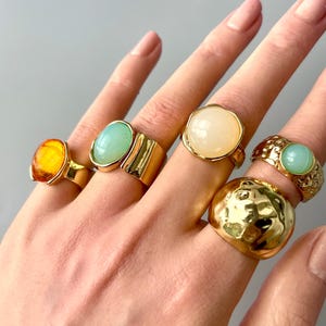 May include: A hand displays six gold-toned rings with various gemstone settings. The rings feature amber, light green, and cream-colored stones. One ring has a textured, hammered design. The rings are in different shapes and sizes.