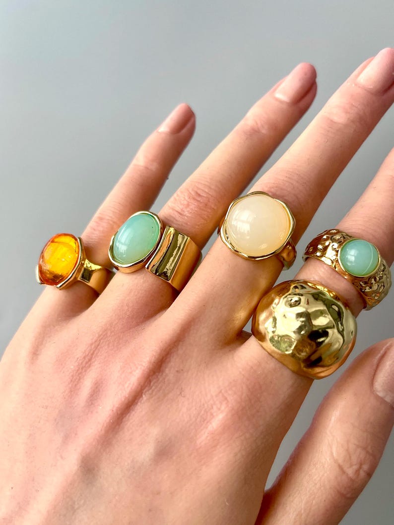 May include: A hand displays six gold-toned rings with various gemstone settings. The rings feature amber, light green, and cream-colored stones. One ring has a textured, hammered design. The rings are in different shapes and sizes.