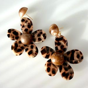 May include: A pair of flower-shaped earrings with a tortoiseshell pattern. Each earring features a large, five-petal flower design with a brown and black spotted pattern. The earrings have a small, round stud and a matching tortoiseshell bead in the centre.