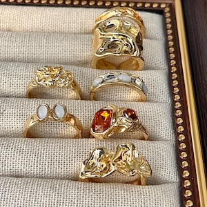 May include: A collection of gold-toned rings displayed in a decorative frame. The rings feature various designs, including textured bands, gemstone accents, and organic shapes. The frame has a brown finish with a beaded border.
