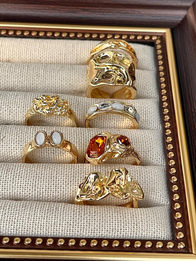 May include: A collection of gold-toned rings displayed in a decorative frame. The rings feature various designs, including textured bands, gemstone accents, and organic shapes. The frame has a brown finish with a beaded border.
