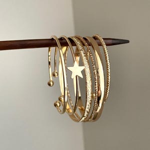 以下が含まれることがあります： 星型のチャームが付いたブレスレットを含む、金色のバングルのコレクション。 バングルは、滑らか、テクスチャード加工、ビーズのデザインなど、さまざまなテクスチャを持っています。 ジュエリーは木の棒に展示されています。