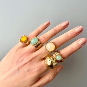 May include: A hand displays six gold-toned rings with various gemstone settings. The rings feature stones in shades of yellow, green, and cream. One ring has a textured, hammered design. The rings are set against a neutral gray background.