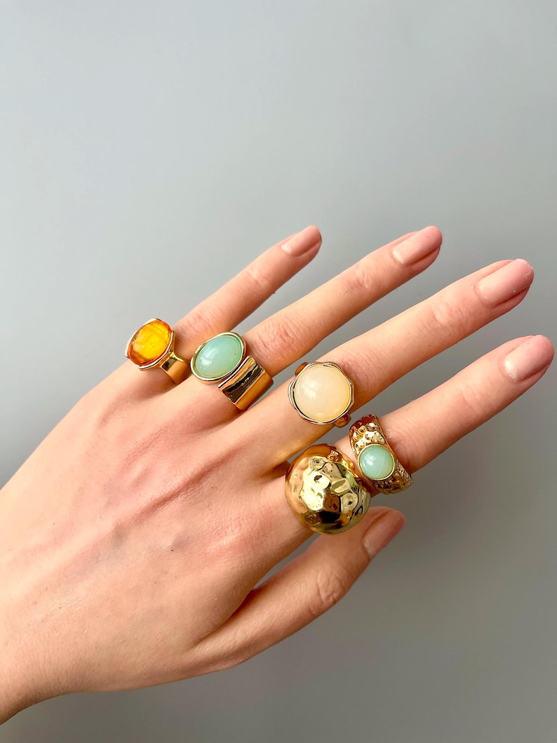 May include: A hand displays six gold-toned rings with various gemstone settings. The rings feature stones in shades of yellow, green, and cream. One ring has a textured, hammered design. The rings are set against a neutral gray background.