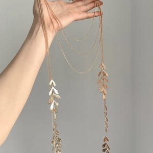 Puede incluir: Un collar dorado con múltiples cadenas finas y colgantes en forma de hojas. El collar se muestra sobre un fondo gris, resaltando su delicada cadena y su diseño detallado. Los colgantes de hojas añaden un toque de elegancia inspirada en la naturaleza.