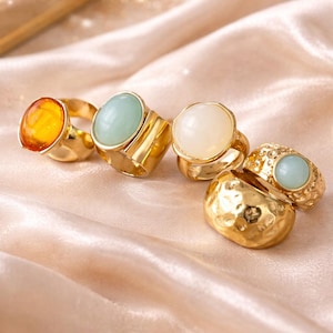 May include: A collection of gold-toned rings with various gemstone settings. The rings feature amber, light green, and white stones. The rings are displayed on a peach-colored satin fabric, creating a luxurious aesthetic.