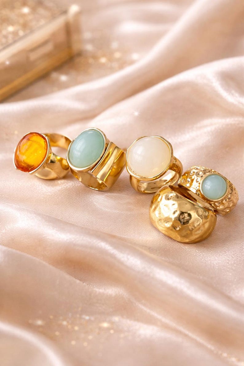May include: A collection of gold-toned rings with various gemstone settings. The rings feature amber, light green, and white stones. The rings are displayed on a peach-colored satin fabric, creating a luxurious aesthetic.