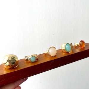 May include: A collection of gold-toned rings with various gemstone settings, including a hammered dome, a light blue stone, a pink stone, a turquoise stone, and an orange stone. The rings are displayed on a wooden surface.