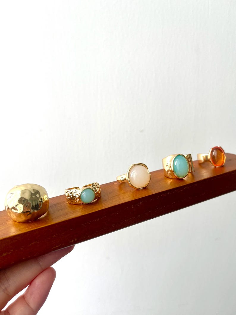 May include: A collection of gold-toned rings with various gemstone settings, including a hammered dome, a light blue stone, a pink stone, a turquoise stone, and an orange stone. The rings are displayed on a wooden surface.