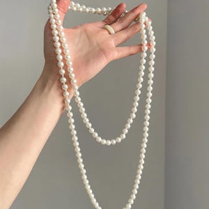 Puede incluir: Un collar largo de perlas blancas se muestra sobre un fondo neutro. El collar está compuesto por muchas perlas pequeñas y redondas. Las perlas son de color blanco cremoso y parecen tener un tamaño uniforme.