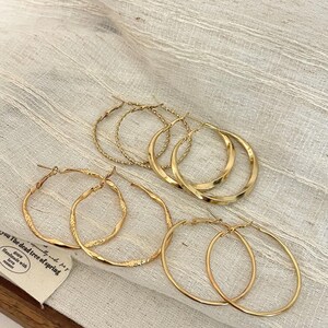 May include: Several pairs of gold-coloured hoop earrings in different designs, including twisted and smooth styles. The earrings are displayed on a light-coloured fabric, showcasing their different textures and shapes. The earrings are a fashion accessory.