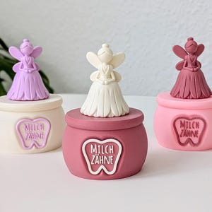 Könnte beinhalten: Drei dekorative Zahnfee-Boxen. Jede Box hat eine Feenfigur oben drauf und die Worte "MILCH ZAHNE" in einem zahnförmigen Design auf der Vorderseite. Die Boxen sind in Pastellfarben: Lavendel, Weiß und Rosa.