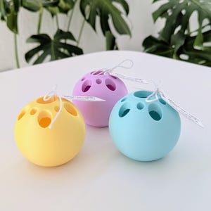 Könnte beinhalten: Drei kugelförmige, perforierte Objekte in Pastellfarben: Gelb, Lavendel und Hellblau. Jede Kugel hat mehrere kreisförmige Öffnungen und ist mit einem weißen Etikett mit einer Schnur versehen. Die Objekte befinden sich auf einer weißen Oberfläche, mit grünem Laub im Hintergrund.