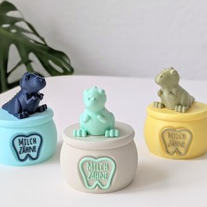 Könnte beinhalten: Drei farbenfrohe Zahnfee-Boxen in Dinosaurierform. Jede Box hat eine andere Farbe: Blau, Mintgrün und Gelb. Die Boxen haben eine runde Form mit Deckel und einer Dinosaurierfigur oben drauf. Der Text "MILCH ZAHNE" befindet sich auf der Vorderseite jeder Box.